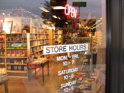 A bookstore entrance with an "OPEN" sign and store hours displayed on the glass door. Shelves filled with books inside.