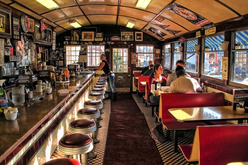 A cozy diner interior with a long counter, red booths, and patrons enjoying meals, surrounded by vintage decor.