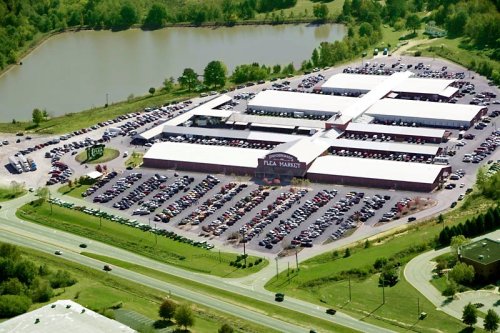 Aerial view of a large flea market with numerous parked cars, surrounded by greenery and a pond nearby.