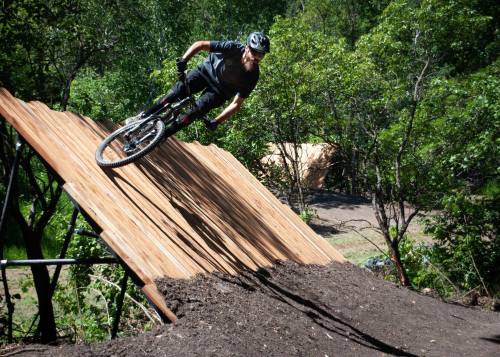 A mountain biker leans into a turn on a wooden ramp surrounded by trees and dirt.