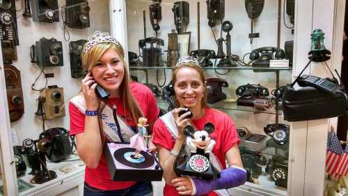 Two women in tiaras hold vintage phones and toys, smiling in front of a display of old telephones.