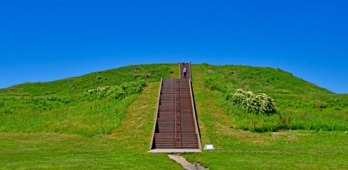 A person walks up a set of stairs leading to a grassy hill under a clear blue sky.