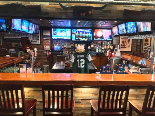 A bar with multiple TVs showing sports, a green jersey hanging, and empty wooden chairs in front of the counter.