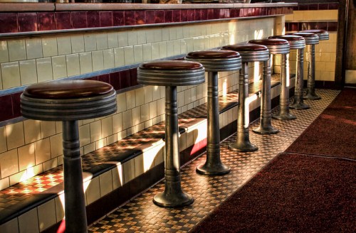 A row of vintage diner stools with red seats, lined up against a tiled counter in warm, soft lighting.