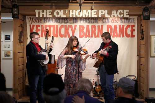 A trio performs on stage at the Kentucky Barn Dance, featuring a banjo, violin, and guitar in a cozy setting.