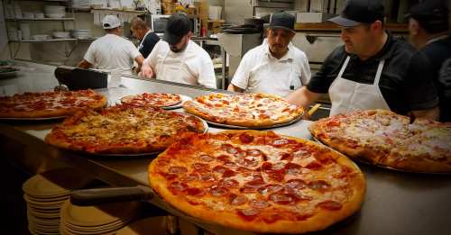A busy kitchen with chefs preparing various pizzas on a counter, showcasing different toppings and styles.