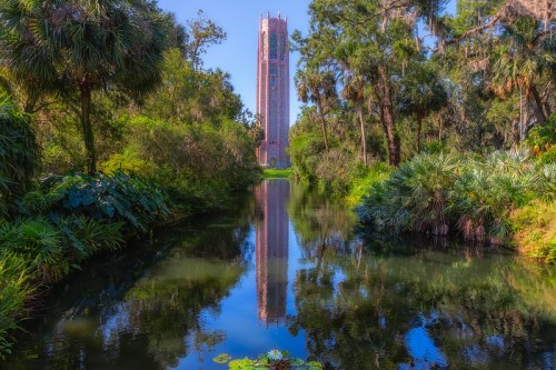 A tall tower reflects in a serene pond, surrounded by lush greenery and palm trees under a clear blue sky.