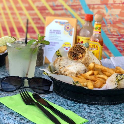 A plate with a burrito, fries, a drink, sunglasses, and condiments on a colorful table.
