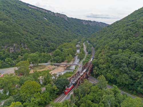A red train crosses a bridge in a lush green valley surrounded by mountains under a cloudy sky.