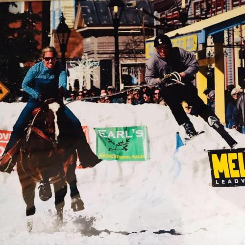 A person on a horse races alongside another person skiing over a snowbank in a festive winter setting.