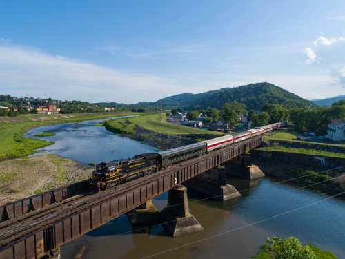 A train crosses a bridge over a river, with green hills and a small town in the background under a clear blue sky.