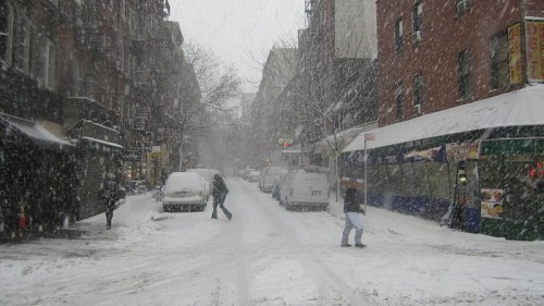 A snowy street scene with heavy snowfall, covered cars, and people walking through the winter weather.