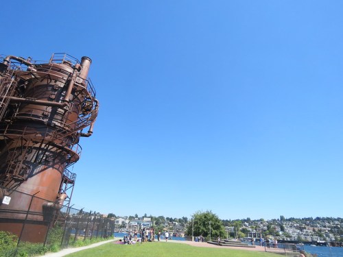 A rusted industrial structure beside a park with people enjoying a sunny day by the water and a city skyline in the background.