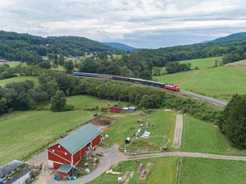 A train travels along a curved track near a red barn and green fields, surrounded by rolling hills and trees.
