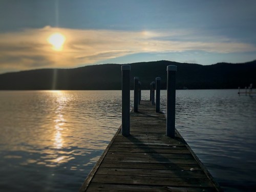 A wooden dock extends into a calm lake, reflecting the sunset and surrounded by mountains.
