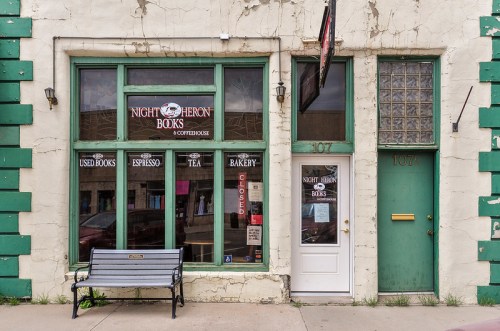 A quaint bookstore with green trim, featuring a bench outside and a sign for used books, espresso, and bakery items.