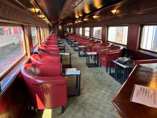 Interior of a vintage train car featuring rows of red leather chairs and small tables, with large windows along the sides.