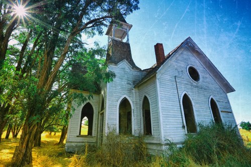 An abandoned white church surrounded by overgrown grass and trees, under a bright blue sky with sun rays.