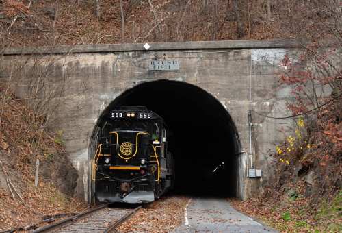 A black train enters a dark tunnel surrounded by autumn foliage and rocky terrain.