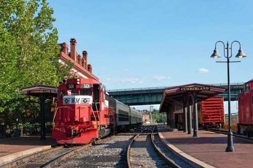 A red train at a station with a platform, surrounded by trees and buildings, under a clear blue sky.