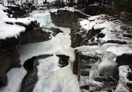 A frozen river flows through rocky terrain, surrounded by snow-covered trees in a winter landscape.