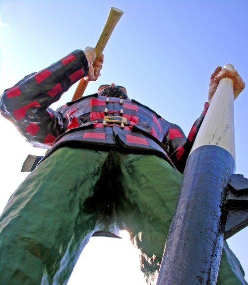 A towering statue of a lumberjack in a plaid shirt, holding two large logs against a clear blue sky.