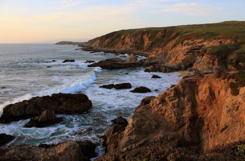 A scenic coastal view with rocky cliffs, gentle waves, and a sunset sky over the ocean.