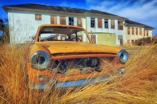 An abandoned orange car sits in tall grass in front of a dilapidated building with boarded windows.