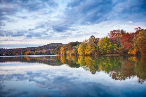 A serene lake reflects colorful autumn trees under a cloudy sky, creating a peaceful natural landscape.