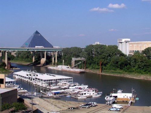 A river scene featuring boats docked, a bridge overhead, and a pyramid-shaped building in the background under a clear sky.