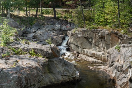 A serene rocky landscape with a small waterfall flowing into a calm stream, surrounded by trees and greenery.