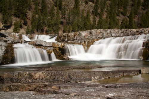 A serene waterfall cascades over rocky ledges, surrounded by lush green trees and a tranquil pool below.