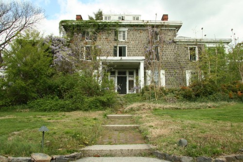 A large, overgrown stone house with a front porch, surrounded by greenery and trees under a cloudy sky.