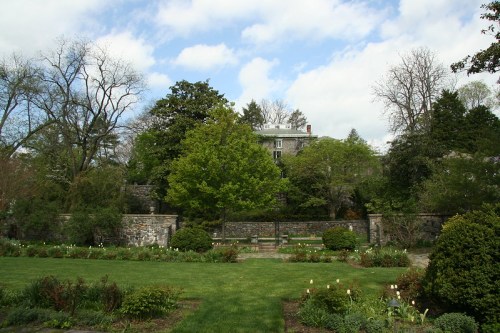A lush garden with green grass, trees, and a stone wall, set against a cloudy sky and a distant building.