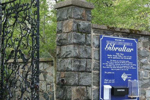 A stone gate with a blue sign detailing visiting hours and donation information for Gibraltar. Lush greenery surrounds it.