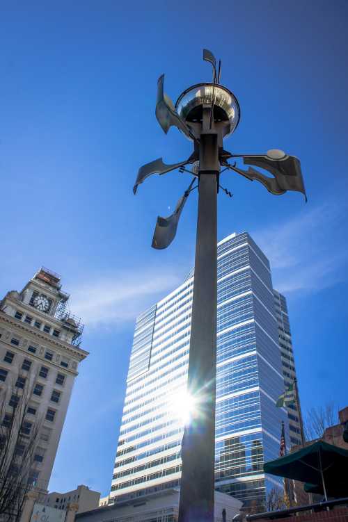 A tall sculpture with metal blades stands in front of a modern skyscraper under a clear blue sky.