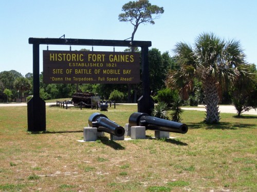 Sign for Historic Fort Gaines with cannons in front, surrounded by grass and palm trees, marking the Battle of Mobile Bay.