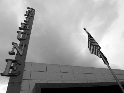 A black and white image of a building sign reading "Tribune" with an American flag waving against a cloudy sky.