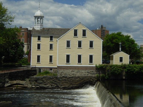 Historic yellow mill building beside a river with a small waterfall, under a partly cloudy sky.
