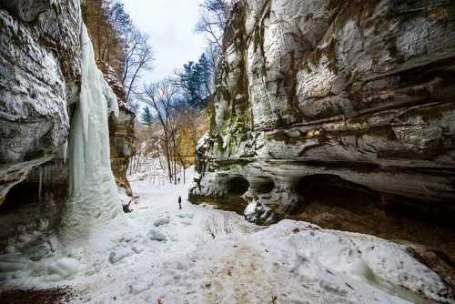 A person stands in a snowy canyon surrounded by icy cliffs and frozen waterfalls under a cloudy sky.