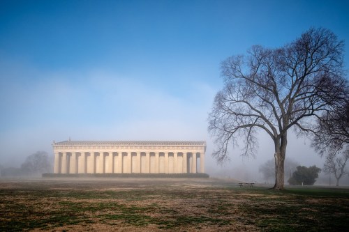 A foggy landscape featuring a classical building with columns and a bare tree in the foreground.