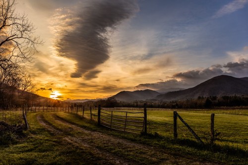 A serene sunset over mountains, with a dirt path leading to a wooden gate and a grassy field.