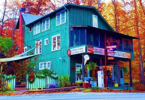 A colorful, vintage grocery store with a green exterior, surrounded by autumn foliage and a welcoming open sign.