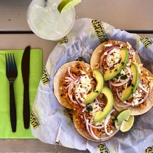 Three tacos filled with shredded chicken, topped with avocado, onions, and cheese, served with a drink and utensils.