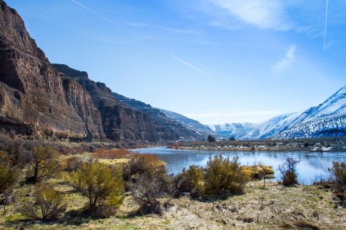 A serene river flows between snow-capped mountains and rocky cliffs under a clear blue sky.