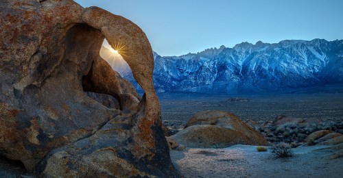 A rocky arch frames a stunning view of snow-capped mountains at sunrise, casting warm light across the landscape.