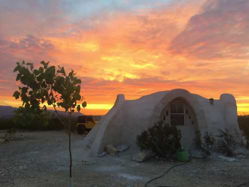 A unique adobe-style structure against a vibrant sunset, with a small tree and desert landscape in the foreground.