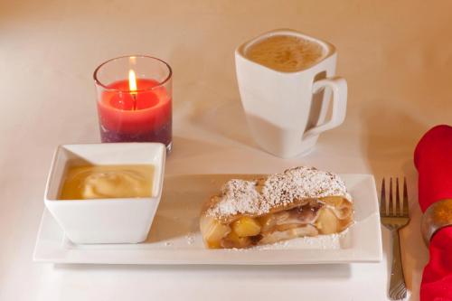 A plate with apple strudel dusted with powdered sugar, a cup of coffee, a candle, and a bowl of cream sauce.