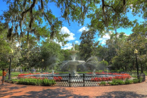 A scenic park featuring a fountain surrounded by colorful flowers and lush trees under a bright blue sky.
