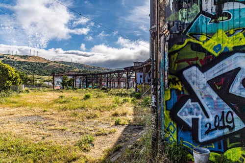 A vibrant graffiti-covered wall beside an overgrown field and a dilapidated structure under a cloudy sky.